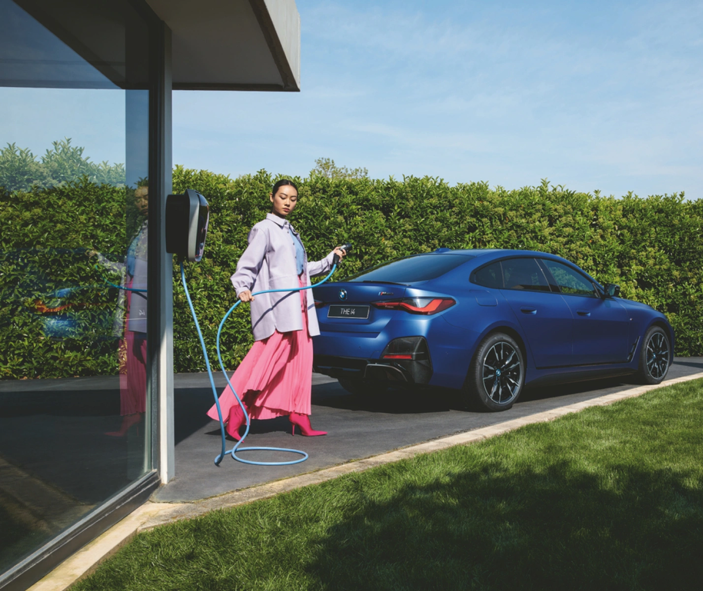Person charging a blue electric car at an outdoor charging station next to a modern house with glass walls.